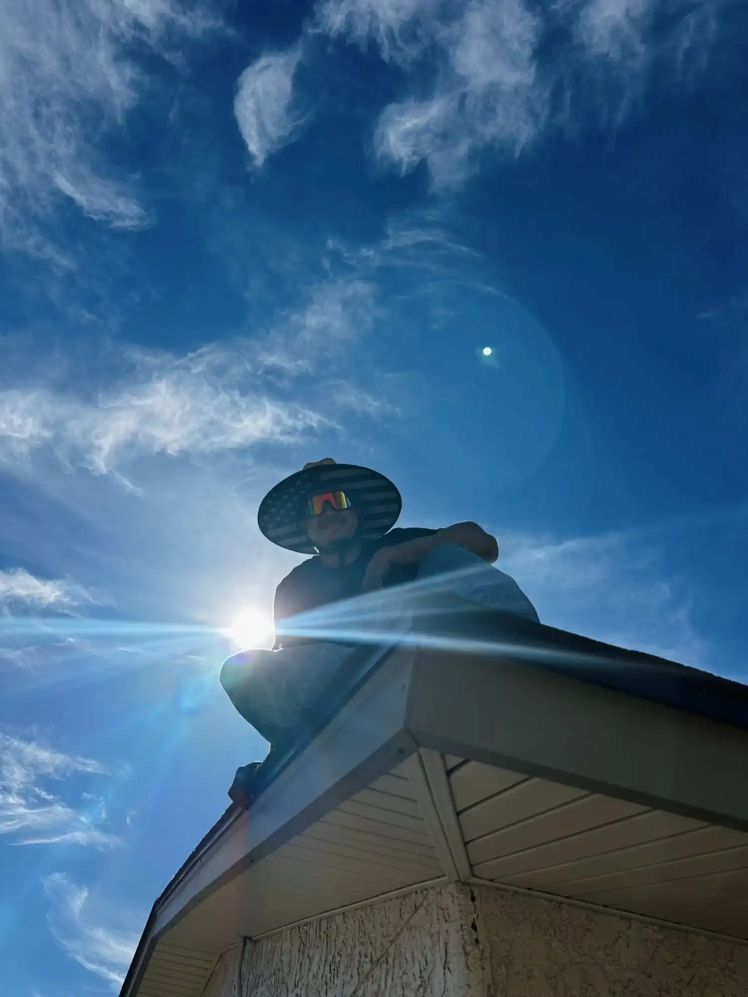 A DC AC Arizona technician in a wide-brim hat sitting on a roof edge, silhouetted against the sun.