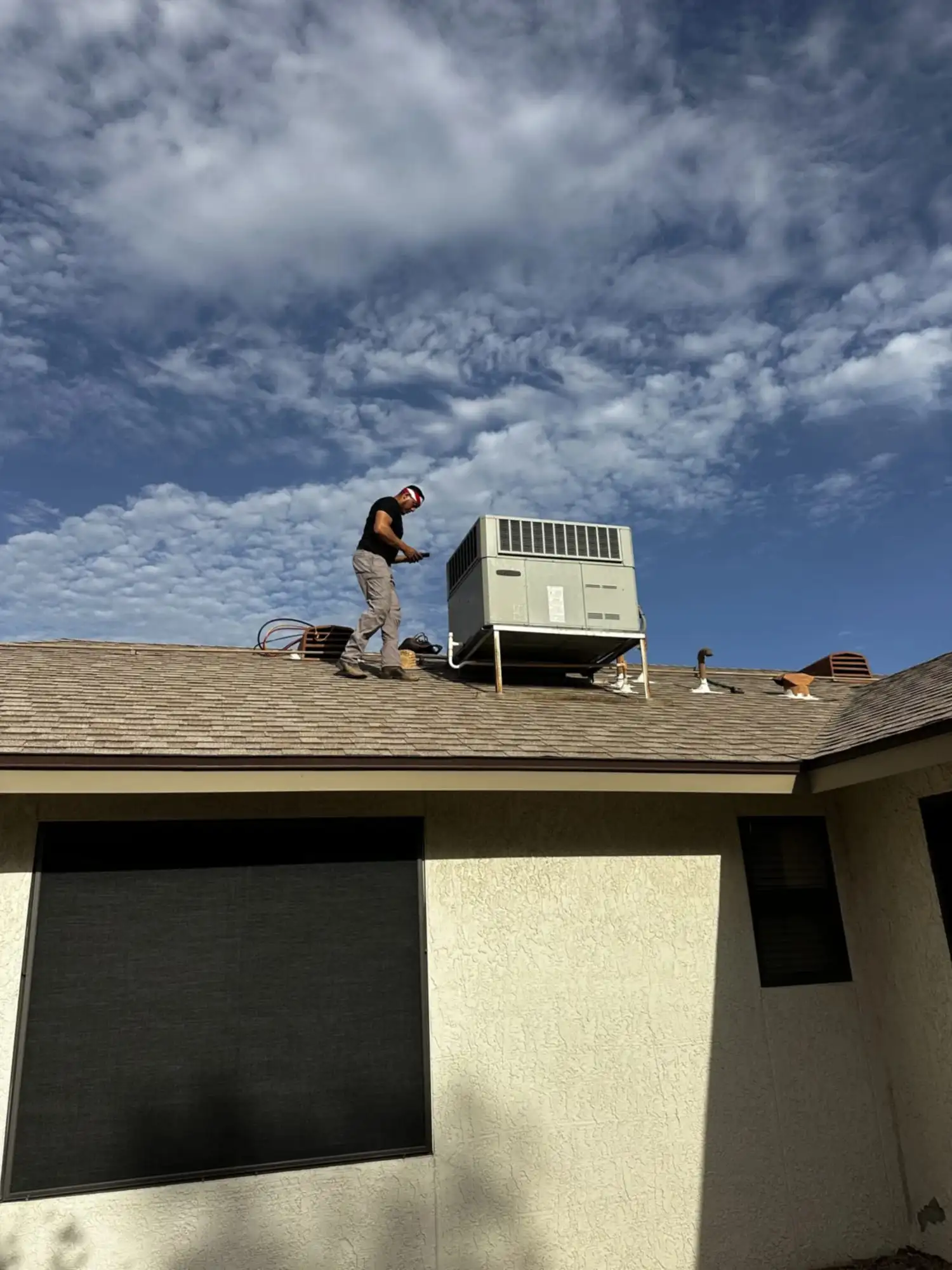 A DC AC Arizona technician standing on a roof next to a gray rooftop package unit, under a cloud-streaked sky.
